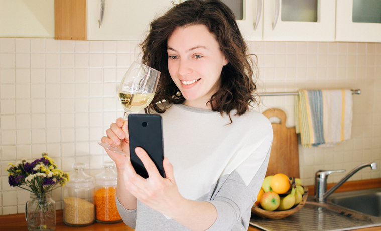 Smiling young woman making video call with smartphone at kitchen. Best friends drinking white wine and toasting online on a video call during quarantine. Stay home save lives from covid-19. Isolation