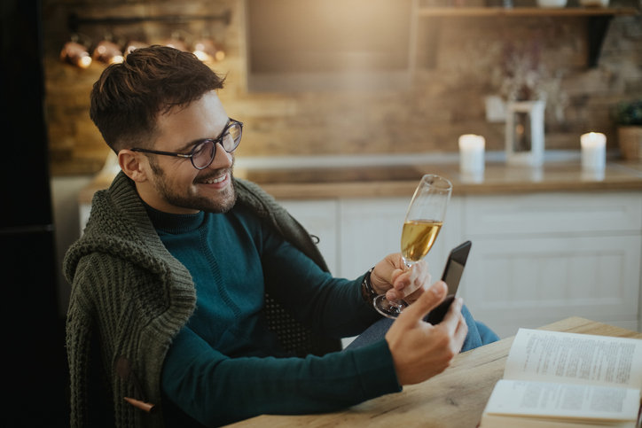 Young happy man using smart phone and toasting with Champagne during video call at home.