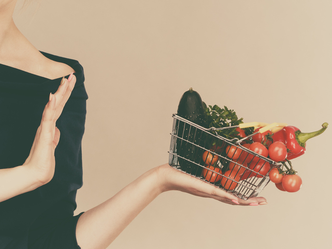 Adult woman do not like to eat vegetables, healthy food, vegetarian products. Female holding small shopping basket with green red vegetables, stop gesture, on grey