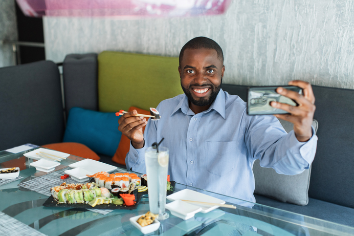 Excited smiling young African business man doing selfie shot or video call on mobile phone, holding chopsticks with sushi roll, having traditional oriental lunch at restaurant