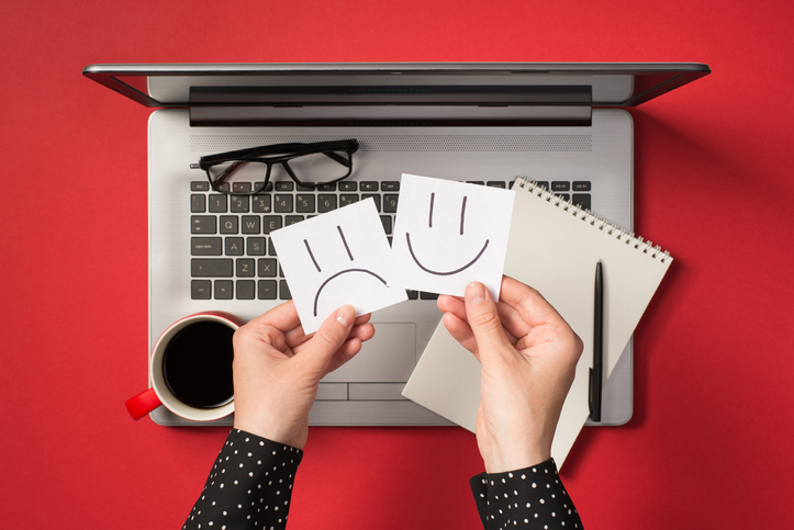First person top view photo of female hands holding two sticker notes with drawn happy and sad faces over open grey laptop glasses and red cup of drink on isolated red background