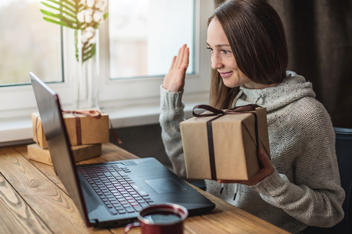 A girl in a sweater with a gift box in her hands is sitting in front of a computer screen and waving her hand. Concept of distance gifts and surprises and communicate through video link.