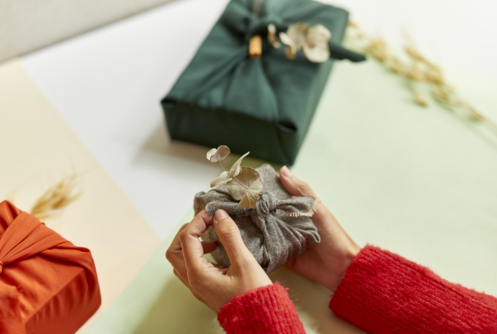 Female hands holding gift wrapped in fabric on a table with other presents, Christmas and New year minimalist concept