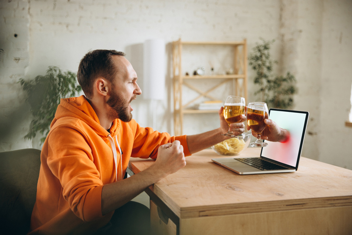 Clinking. Young man drinking beer during meeting friends on virtual video call. Distance online meeting, chat together on laptop at home. Concept of remote safe meetings and entertainment.