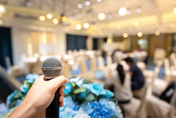 A speaker's hand is holding wireless microphone, prepare for speaking in the education seminar or business townhall conference on the stage. Close-up and mic and hall room as blurred background.