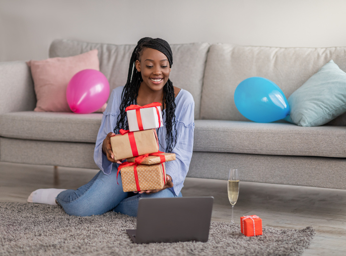 Happy young black woman with gift boxes smiling at laptop screen, sitting on floor by sofa at home, having online party, International Womens Day celebration during COVID-19 pandemic, copy space