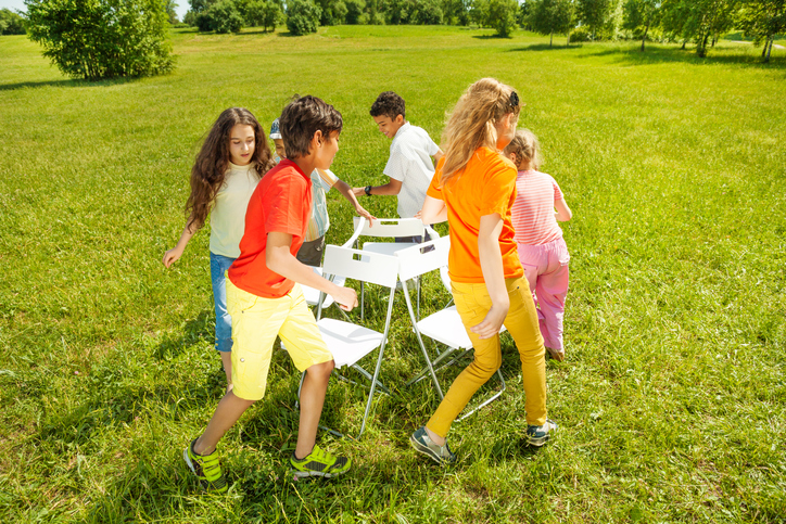 Kids run around chairs playing a game outside in summer period
